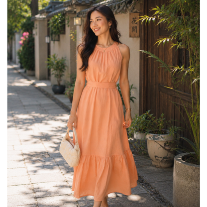 A woman wearing a sleeveless peach-orange tiered midi dress walks down a sunlit stone pathway beside traditional buildings and potted plants, holding a cream handbag.